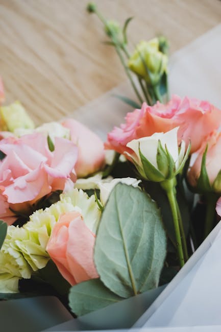 A close-up of a floral bouquet featuring soft pink and pale yellow eustoma (lisianthus) flowers with layered, ruffled petals, accompanied by green foliage and leaves. The flowers are arranged in a del