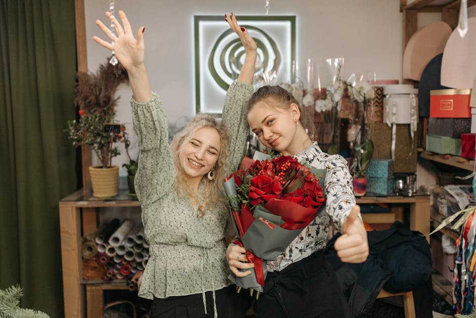 Two young women inside a flower shop, smiling cheerfully while holding a large bouquet of fresh, vibrant red roses wrapped in gray paper with red ribbon. The woman on the right is giving a thumbs-up g