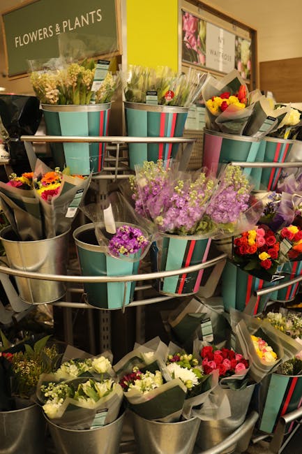 A display of various floral bouquets arranged in metallic buckets on a multi-tiered stand inside a florist shop. The arrangements include a mix of fresh flowers such as purple statice, yellow and red 