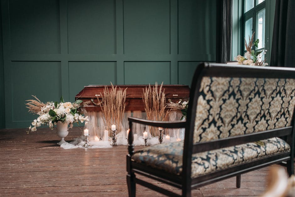 An indoor setting featuring a wooden funeral urn decorated with dried pampas grass and white floral arrangements in elegant white vases. Surrounding the urn are small lit candles, adding a solemn ambi