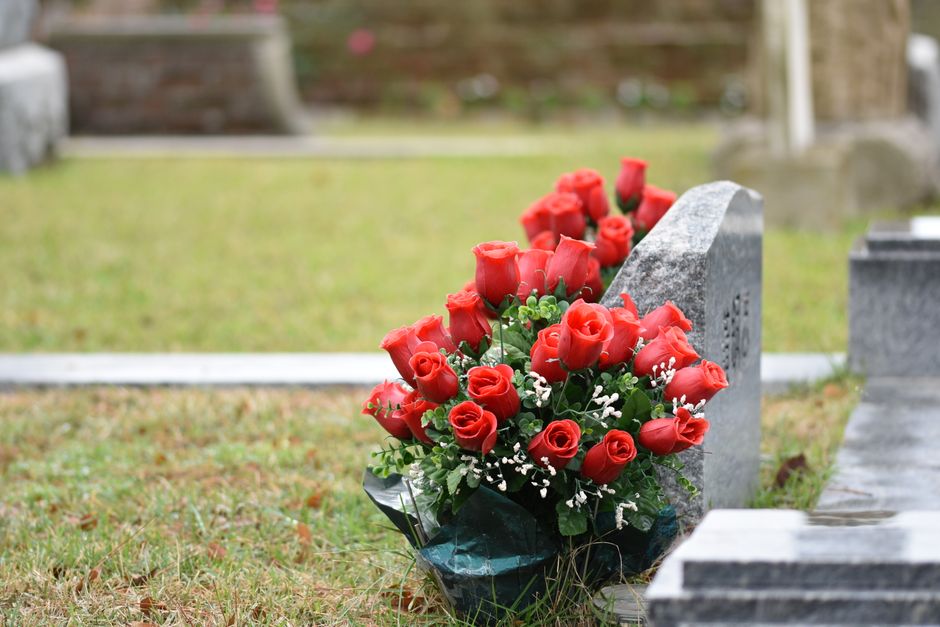 A bouquet of fresh, vibrant red roses with tightly packed petals and glossy green leaves, arranged in a lush cluster and wrapped in dark green paper, rests against a gray stone gravestone in a well-ma