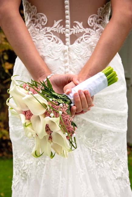 A bride in a sleeveless lace wedding gown holding a bouquet of fresh white calla lilies mixed with pink baby's breath and green foliage, wrapped in a white ribbon, with her hands gently clasped around