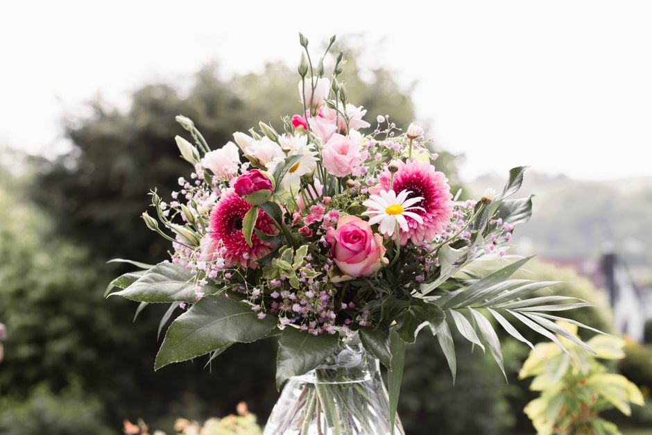 A vibrant bouquet of fresh flowers arranged in a clear glass vase, featuring pink roses, white daisies with yellow centers, and pink gerbera daisies, complemented by eucalyptus and palm leaves. The bo