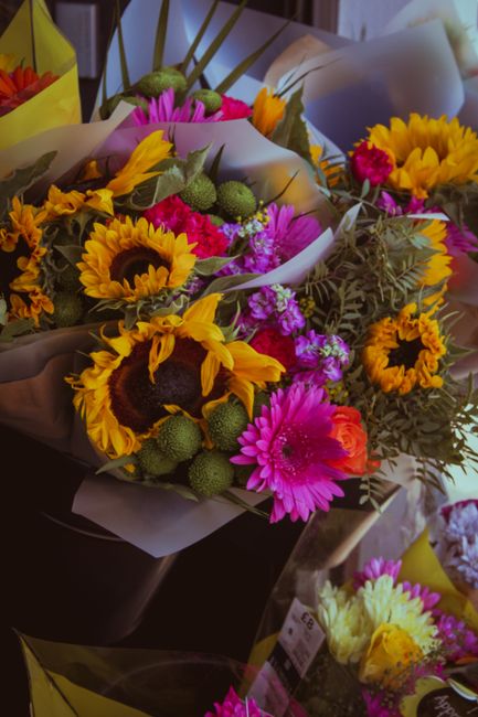A vibrant floral bouquet featuring large yellow sunflowers with dark centers, pink gerbera daisies, small purple and pink flowers, and green foliage arranged in white wrapping paper. The bouquet is pl